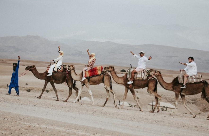 Guests waving from camelback with the Atlas Mountains behind